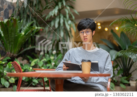 Glasses asian man reading book writing a note with coffee cup on table while sitting in garden cafe. 138302211
