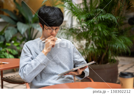 Glasses asian man holding pen thinking and looking at tablet with coffee on table sitting in garden. 138302212
