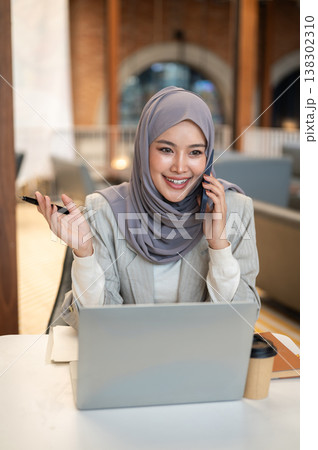 Pretty asian muslim woman talking on phone in front of laptop aside book while sitting at cafe table 138302310