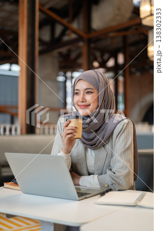 Pretty asian muslim woman student drinking coffee over laptop aside book while sitting at cafe table Pretty asian muslim woman student drinking coffee over laptop aside book while sitting at cafe table 138302333