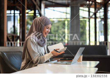 Asian muslim woman holding book talking explaining on video call via laptop while sits at cafe table Asian muslim woman holding book talking explaining on video call via laptop while sits at cafe table 138302338