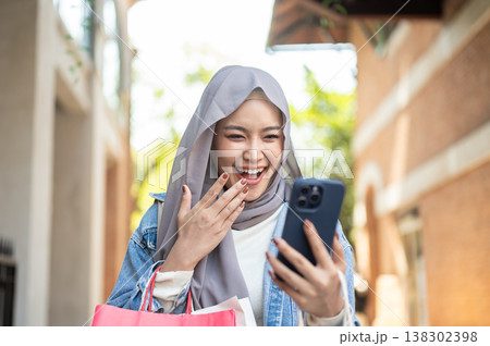 Happy asian muslim woman tourist looking at phone hand covering mouth as walking shopping in market. Happy asian muslim woman tourist looking at phone hand covering mouth as walking shopping in market. 138302398