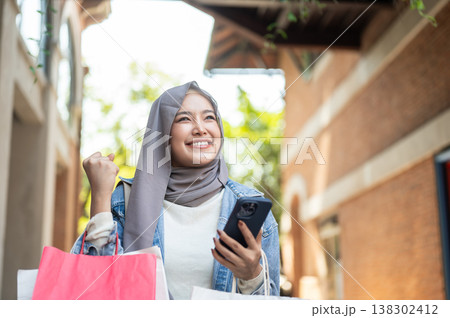 Happy asian muslim woman tourist holding phone carrying shopping bag making a fist walking in market Happy asian muslim woman tourist holding phone carrying shopping bag making a fist walking in market 138302412