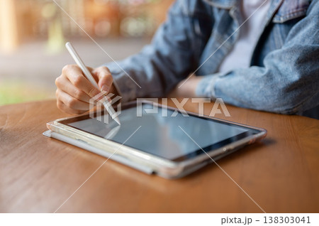 Close up of woman's hand holding pen writing drawing on tablet at wooden table while sitting in cafe Close up of woman's hand holding pen writing drawing on tablet at wooden table while sitting in cafe 138303041