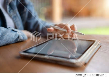 Close up of a woman's hand holding pen touching on tablet at wooden table while sitting in the cafe. 138303042