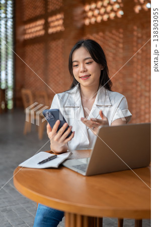 Pretty asian woman holding and looking at phone over book and laptop sitting at wooden table in cafe Pretty asian woman holding and looking at phone over book and laptop sitting at wooden table in cafe 138303055