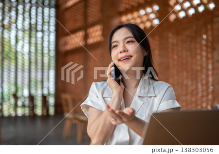 Pretty asian woman holding phone talking on call over laptop while sitting in wooden pavilion cafe. 138303056