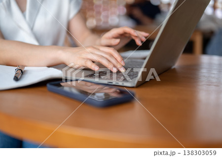 Close up of woman's hand typing on laptop aside book and phone while sitting at wooden table in cafe 138303059