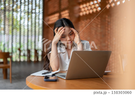 Stressed asian woman holding head looking at laptop aside book on wooden table while sitting in cafe Stressed asian woman holding head looking at laptop aside book on wooden table while sitting in cafe 138303060