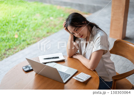 Asian woman holding head looking at laptop aside calculator in troubled while sitting in garden cafe 138303064