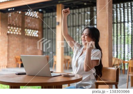 Happy asian woman looking at laptop raising a fist in excited while sitting at wooden table in cafe. 138303066