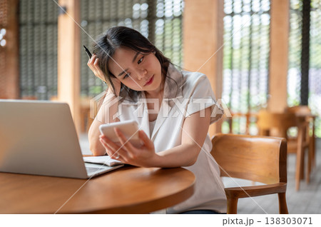 Confused asian woman holding pen looking at calculator aside laptop while sitting at table in cafe. Confused asian woman holding pen looking at calculator aside laptop while sitting at table in cafe. 138303071