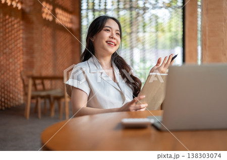 Smiling asian woman holding pen and book thinking aside laptop on wooden table while sitting in cafe Smiling asian woman holding pen and book thinking aside laptop on wooden table while sitting in cafe 138303074