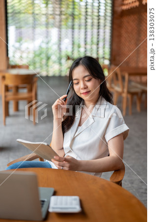 Pretty asian woman holding pen looking at book thinking aside laptop on table while sitting in cafe. Pretty asian woman holding pen looking at book thinking aside laptop on table while sitting in cafe. 138303075