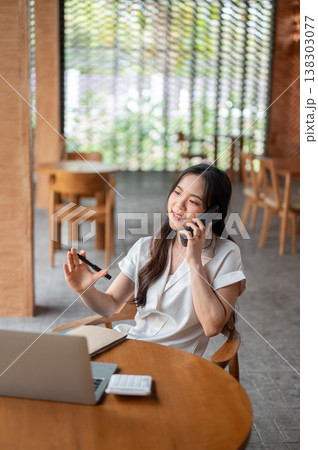 Pretty asian woman holding a pen talking on phone call aside laptop on table while sitting in cafe. Pretty asian woman holding a pen talking on phone call aside laptop on table while sitting in cafe. 138303077