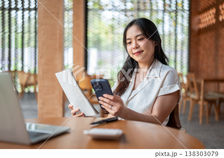 Pretty asian woman holding book looking at phone aside laptop while sitting at wooden table in cafe. Pretty asian woman holding book looking at phone aside laptop while sitting at wooden table in cafe. 138303079