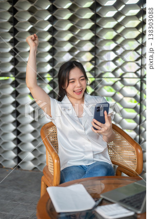 Happy asian woman looking at phone raising a fist in excited sitting on wooden armchair in the cafe Happy asian woman looking at phone raising a fist in excited sitting on wooden armchair in the cafe 138303088