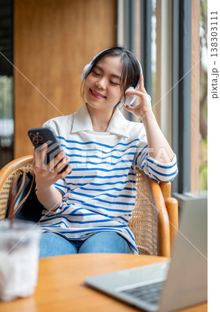 Happy asian woman enjoy listening to music in headphones sitting with laptop at wooden table in cafe Happy asian woman enjoy listening to music in headphones sitting with laptop at wooden table in cafe 138303111