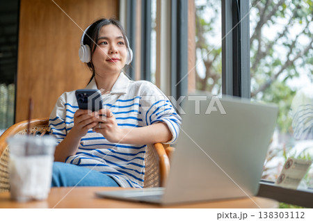 Happy asian woman listening to music holding a phone sitting with laptop at table by window in cafe. Happy asian woman listening to music holding a phone sitting with laptop at table by window in cafe. 138303112
