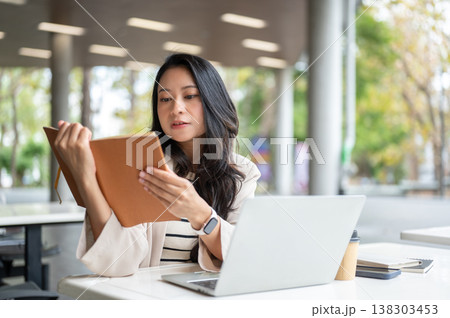Asian woman reading a book aside laptop and coffee cup while sitting at table in university building 138303453