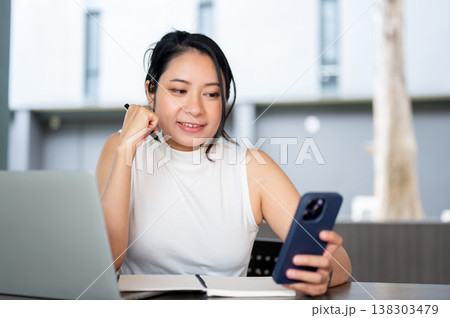 Mature asian working woman holding pen looking at phone over book and laptop sitting at wooden table Mature asian working woman holding pen looking at phone over book and laptop sitting at wooden table 138303479
