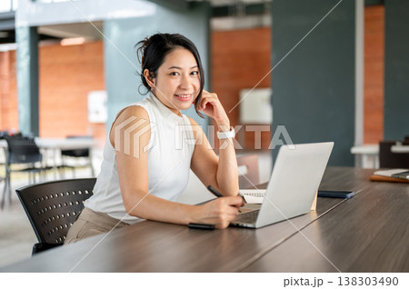 Pretty mature asian woman holding pen over laptop aside book while sitting at wooden table in cafe. 138303490