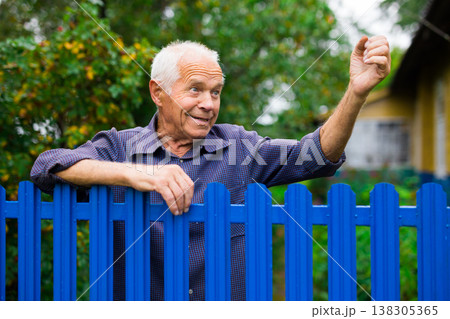 Old elderly farmer stands at fence of his country house and threatens someone with his fist Old elderly farmer stands at fence of his country house and threatens someone with his fist 138305365