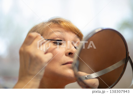 Middle aged woman applying mascara to her eyelashes looking into a mirror. Mature lady performing daily morning makeup routine. Beauty self care ritual for skin and face appearance. Middle aged woman applying mascara to her eyelashes looking into a mirror. Mature lady performing daily morning makeup routine. Beauty self care ritual for skin and face appearance. 138307943