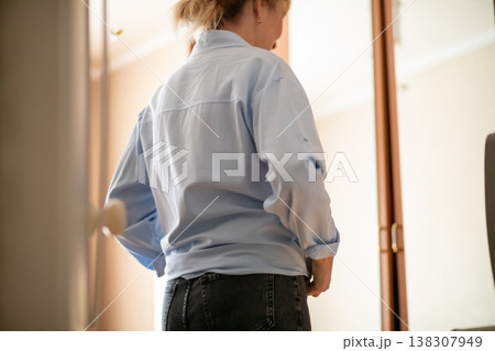 Middle aged woman looking in mirror at home. Caucasian lady standing in casual blue shirt and denim jeans. Routine interior scene of mature female self reflection and morning dressing. Middle aged woman looking in mirror at home. Caucasian lady standing in casual blue shirt and denim jeans. Routine interior scene of mature female self reflection and morning dressing. 138307949