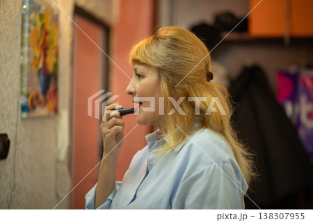 Middle aged woman applying lipstick in front of mirror. 57 years old female beauty routine at home. Daily makeup process and skin care concept. Mature lady preparing for work or event. 138307955