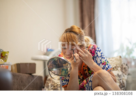 Middle aged woman applying eyeliner looking at small mirror in living room. Daily morning beauty makeup routine of 57 year old lady. Self care and aging gracefully concept at home. Middle aged woman applying eyeliner looking at small mirror in living room. Daily morning beauty makeup routine of 57 year old lady. Self care and aging gracefully concept at home. 138308108