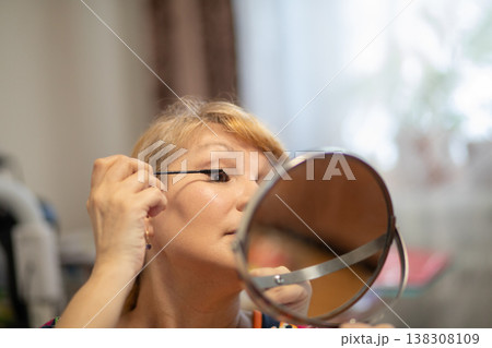 Middle aged woman applying mascara to her eyelashes looking in small mirror. Daily beauty routine of 57 years old female. Self care process at home. Caucasian lady doing makeup. 138308109