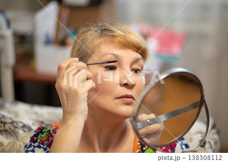 Middle aged woman applying mascara with mirror. Mature female doing makeup at home. Beauty routine for older person to enhance lashes. skincare and aging concept for cosmetic industry. 138308112