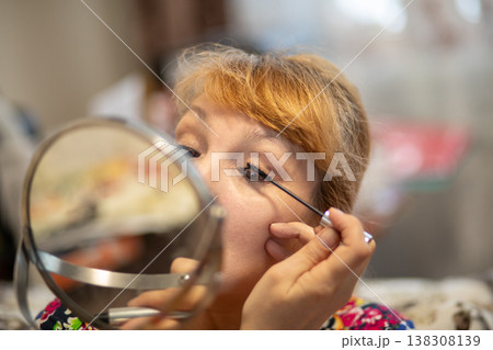 Middle aged woman applying mascara to her eyelashes looking into a mirror. Mature lady practicing beauty makeup routine at home. Self care, cosmetic lifestyle and skincare concept. Middle aged woman applying mascara to her eyelashes looking into a mirror. Mature lady practicing beauty makeup routine at home. Self care, cosmetic lifestyle and skincare concept. 138308139