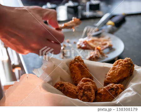 Man eating deep-fried chicken wings from a paper basket. Crispy appetizer served at home. Casual meal, hungry person enjoying fast food snack. Delicious culinary experience and diet concept. 138308179