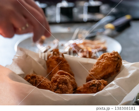 Man eating deep-fried chicken wings from a paper basket. Crispy appetizer served at home. Casual meal, hungry person enjoying fast food snack. Delicious culinary experience and diet concept. 138308181