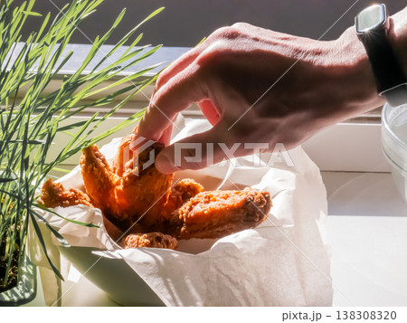 Man hand grabbing crispy fried chicken wing from bowl. Fast food snack served in paper. Homemade meal preparation, culinary concept. Greasy appetizer, protein dish, kitchen interior. Man hand grabbing crispy fried chicken wing from bowl. Fast food snack served in paper. Homemade meal preparation, culinary concept. Greasy appetizer, protein dish, kitchen interior. 138308320