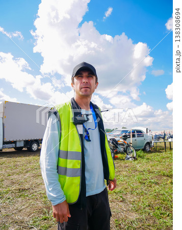 Safety officer man wearing yellow reflective vest and cap standing outdoors at event site. Professional marshal managing logistics and security during outdoor sport competition. Safety officer man wearing yellow reflective vest and cap standing outdoors at event site. Professional marshal managing logistics and security during outdoor sport competition. 138308694