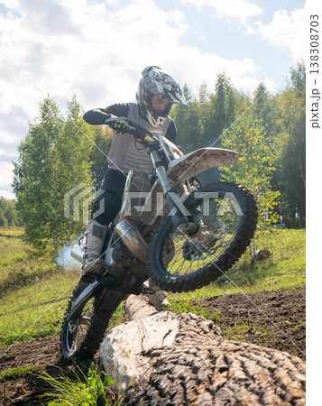 Motocross rider performing wheelie over fallen tree log in forest. Man racing dirt bike on off-road terrain. Extreme sports activity, speed, adrenaline, skill and outdoor adventure ride. Motocross rider performing wheelie over fallen tree log in forest. Man racing dirt bike on off-road terrain. Extreme sports activity, speed, adrenaline, skill and outdoor adventure ride. 138308703