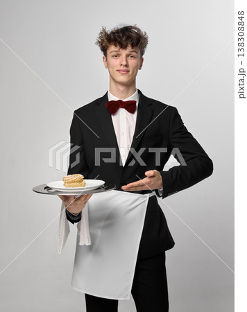 Smiling waiter holding dessert and gesturing toward plate in clean studio portrait. Smiling waiter holding dessert and gesturing toward plate in clean studio portrait. 138308848