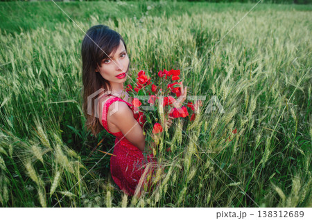 A horizontal high-angle shot of a beautiful young woman with long dark hair and red lipstick, wearing a red patterned dress and holding a bouquet of red poppies while sitting among tall green wheat. 138312689