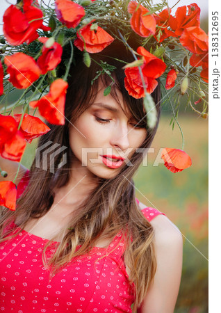A vertical close-up portrait of a beautiful young woman with long dark hair, wearing a red patterned dress and a large wreath of red poppies, 138312695