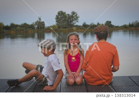 A group of young children sit on a wooden pier by a lake. The young children look toward the future. The concept of summer, holidays, and a carefree childhood. Summer and childhood concept. Children A group of young children sit on a wooden pier by a lake. The young children look toward the future. The concept of summer, holidays, and a carefree childhood. Summer and childhood concept. Children 138313014