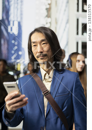 Portrait of asian businessman scrolling on phone among pedestrians in a downtown street, showing multicultural crowd, blurred motion and realistic routine of active urban environment. Portrait of asian businessman scrolling on phone among pedestrians in a downtown street, showing multicultural crowd, blurred motion and realistic routine of active urban environment. 138313570
