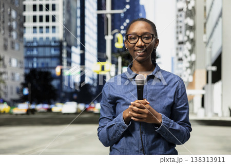 Portrait of smiling street interviewer filming news segment in city center using mic. Happy media professional on city sidewalk using microphone to produce reportage for local television station 138313911