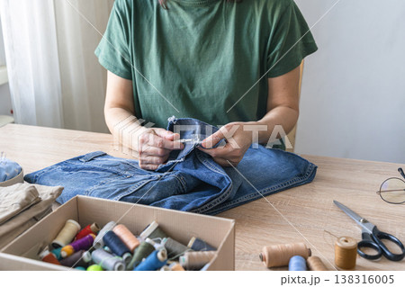 Woman mending blue denim pants by hand, focusing on sustainable clothing repair and textile handicraft at home. An adult woman repairing clothes. Reusing things Woman mending blue denim pants by hand, focusing on sustainable clothing repair and textile handicraft at home. An adult woman repairing clothes. Reusing things 138316005