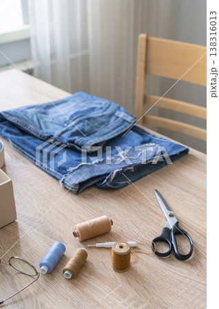 Old denim jeans on a wooden table, surrounded by sewing threads, needles, scissors. Promoting sustainable fashion through craft and repair, upcycling concept 138316013