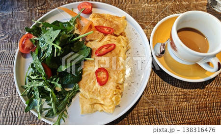 Plate of fluffy omelette garnished with sliced red chili peppers, served alongside fresh salad of arugula and cherry tomatoes, with a cup of coffee on a wooden table Plate of fluffy omelette garnished with sliced red chili peppers, served alongside fresh salad of arugula and cherry tomatoes, with a cup of coffee on a wooden table 138316476