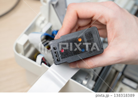 Close-up of a hand removing an ink cartridge from the interior of a printer during maintenance or repair, showing ribbon cable, wiring and mechanical components in a small workspace setting. Close-up of a hand removing an ink cartridge from the interior of a printer during maintenance or repair, showing ribbon cable, wiring and mechanical components in a small workspace setting. 138318389