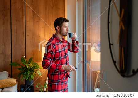 Man in red pajamas drinking coffee near large window at home Man in red pajamas drinking coffee near large window at home 138319307